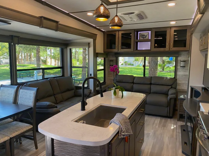 Inside a comfortable RV rental: modern kitchen island with sink, dark sofas, dining table, and large windows showing a green park.. Alliance RV Paradigm 2021
