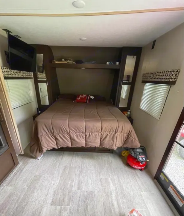 This RV rental features a comfortable bedroom with a brown bed, overhead bunk, TV, and light-tiled floor, perfect for trips.. K-Z Manufacturing Sportsmen 2019