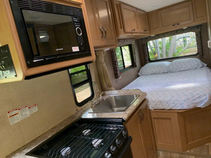 Spacious RV rental interior featuring a full kitchen with microwave, sink, stove, and a cozy bed area.. Thor Four Winds 2019