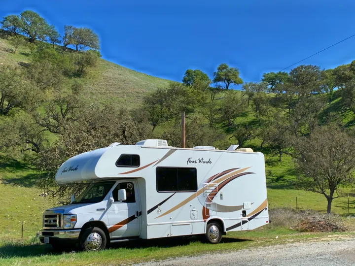 A Four Winds RV rental, a Class C motorhome, parked in a scenic natural setting with a green hillside and scattered trees.. Thor Four Winds 2019