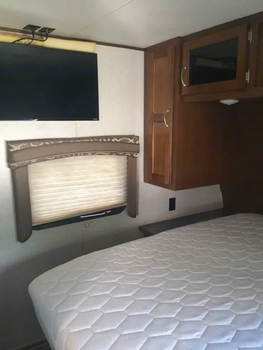 Cozy RV rental bedroom with white quilted bed, wall-mounted TV, and wood storage cabinets. Includes window and exit sign.. Jayco Jay Flight 2018