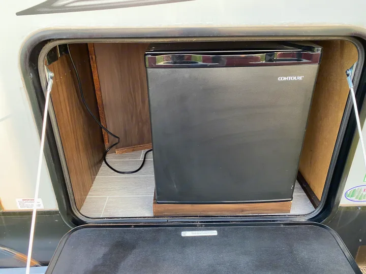 RV rental's exterior storage bay with a black Contoure mini-fridge, ideal for an outdoor kitchen setup.. Forest River Cherokee Alpha Wolf 2020
