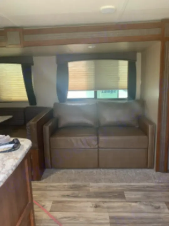 Spacious RV rental interior featuring a brown sofa, wood-look flooring, and large windows with blinds for comfortable living.. Keystone Passport 2017