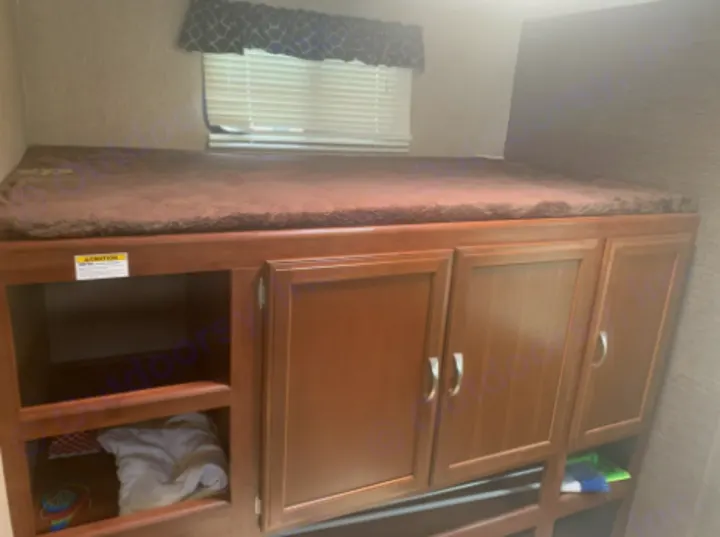 RV rental interior: cozy bunk bed above wooden storage with open shelves and closed cabinets, ideal for family trips.. Keystone Passport 2017