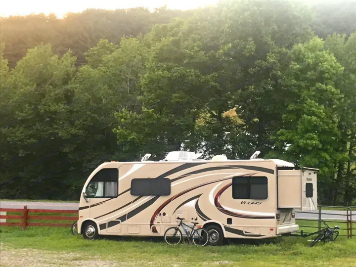 A beige RV rental, model Vegas, with extended slide-out and bikes, parked in a grassy area amidst green trees and a fence.. Thor Vegas 2016