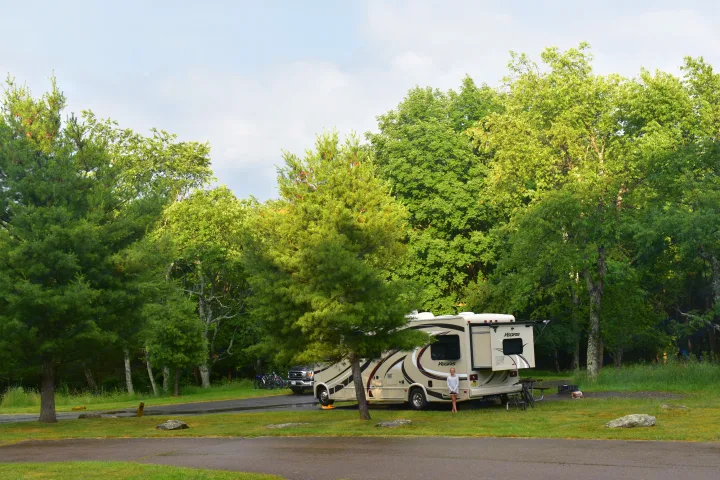 An RV rental, a Vegas motorhome, is parked at a lush, treed campsite with a young girl, suggesting a relaxing family camping trip.. Thor Vegas 2016