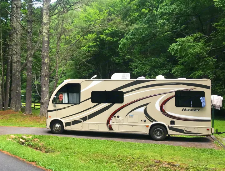 A beige Vegas RV rental is parked in a green, wooded campground, with a person's face visible in the driver's window.. Thor Vegas 2016