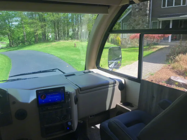 Inside an RV rental cockpit, view out to a paved driveway, green landscape, and a house. The dashboard screen displays "Carry on Wayward Son" by Kansas.. Thor Vegas 2016