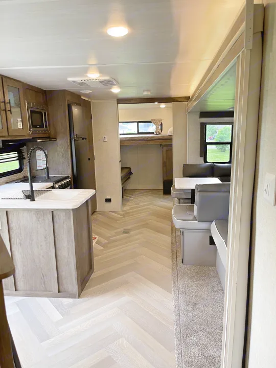 This RV rental interior features a modern kitchen, dining area, and bunk beds with light wood-look herringbone flooring.. Forest River Wildwood 2021