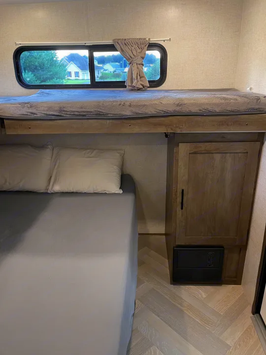 Inside an RV rental: a cozy bedroom featuring a lower bed, upper bunk, window, cabinet, and herringbone flooring.. Forest River Wildwood 2021