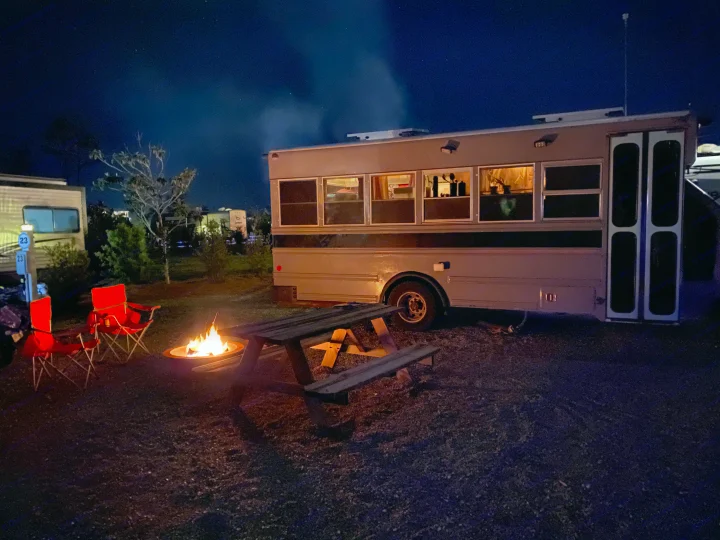 Enjoy a cozy night! This RV rental, a converted bus, features a warm campfire, chairs, and picnic table at a scenic campsite.. Ford E450 2005