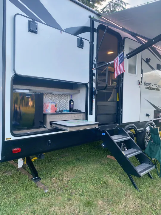 RV rental with side open to reveal an outdoor kitchen, mini-fridge, pull-out table, steps, and lighted awning.. Heartland RVs North Trail 2021