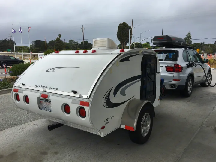 A white Little Guy rv rental teardrop trailer hitched to a silver SUV fueling at a gas station, ready for adventure.. Little Guy Other 2011