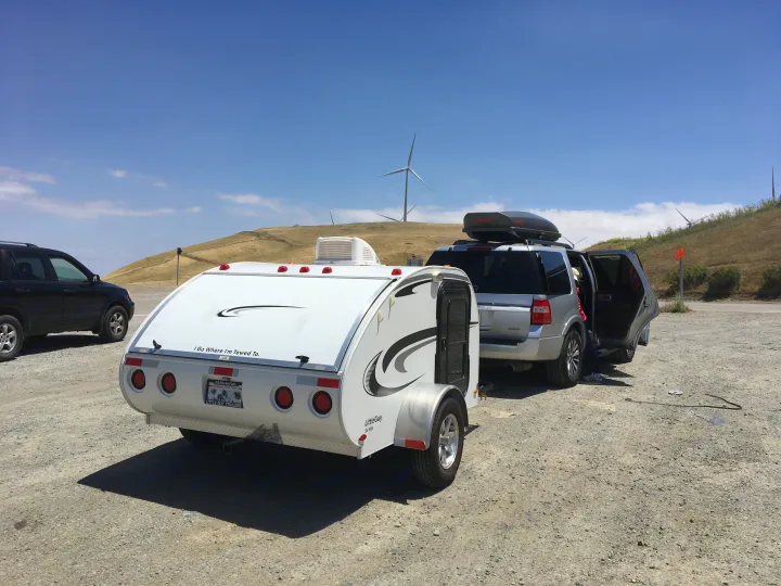 A compact RV rental teardrop trailer is hitched to an SUV on a dirt lot, parked near wind turbines, ready for a road trip.. Little Guy Other 2011