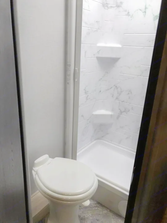 View of a modern RV rental bathroom: a white toilet to the left, and a shower with patterned walls & shelves on the right.. Forest River Sunseeker 2021