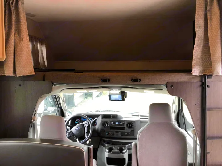 Inside an RV rental, looking forward past the seats to the cockpit with dashboard, steering wheel, and overhead bunk.. Forest River Sunseeker 2021