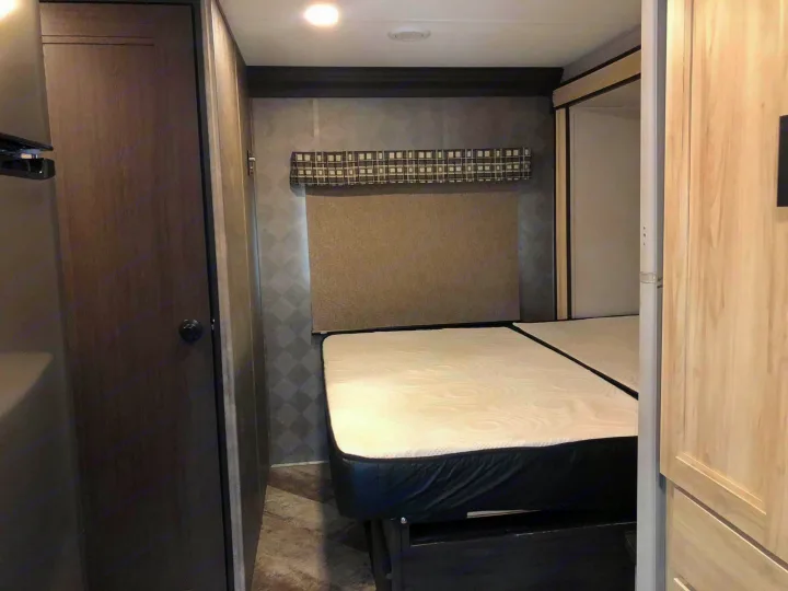 Compact RV rental bedroom featuring a white mattress on a dark base, patterned walls, and wood-paneled doors.. Forest River Sunseeker 2021