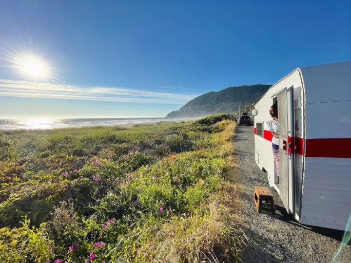 Coastal RV rental. Person looks out from parked RV near stunning beach, ocean, mountains, lush greenery.. Shasta Compact 1968