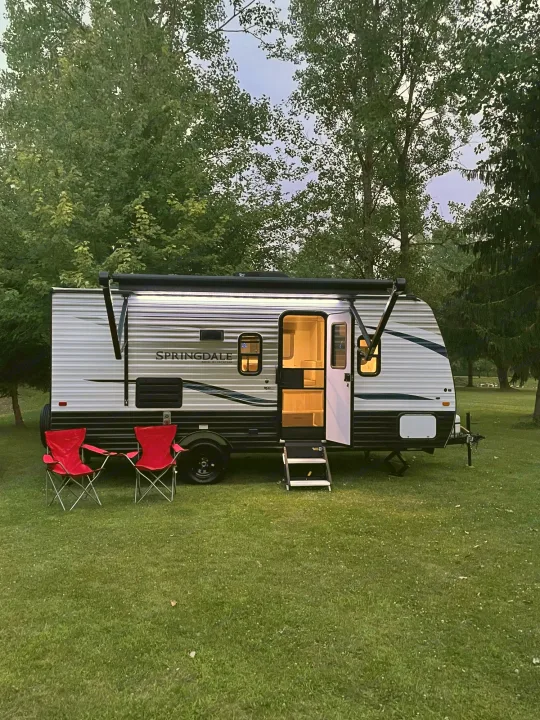 RV rental travel trailer parked at a campsite at dusk. The interior lights are on, and two red camping chairs are set outside.. Keystone Springdale 2021