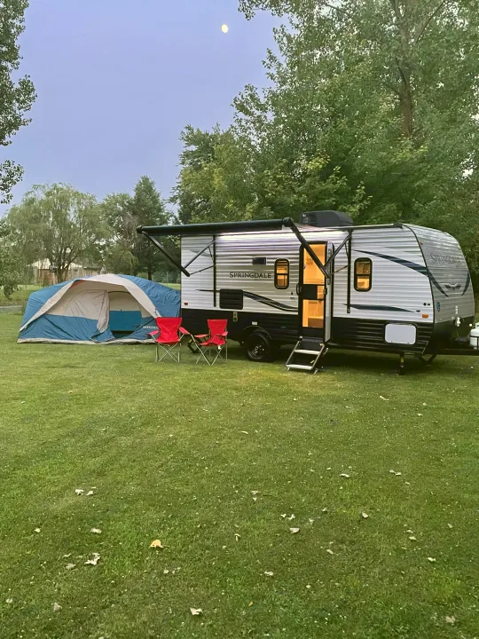 Experience tranquil camping with this RV rental setup: a lighted travel trailer, blue tent, and red chairs on grass at dusk, under a moon.. Keystone Springdale 2021