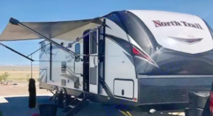 A white & black North Trail travel trailer RV rental, with its awning extended for shade, parked on a sunny day for camping.. North Trail Ultralight 2019