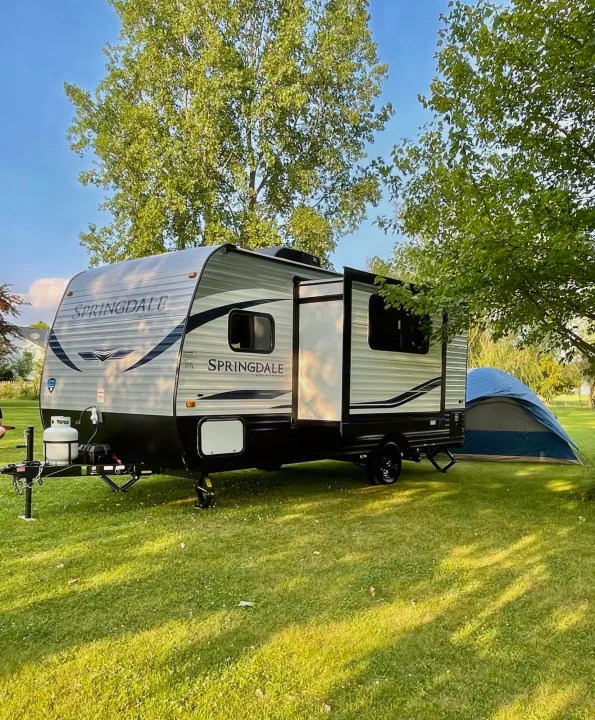 This Springdale Mini RV rental is set up on a grassy lawn with a blue tent behind it, surrounded by trees under clear skies.. Keystone Springdale 2021