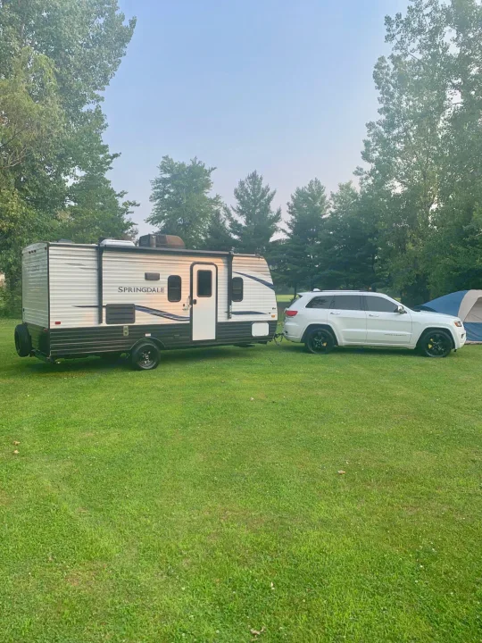 RV rental ready: White SUV towing a Springdale travel trailer at a lush, treed campsite.. Keystone Springdale 2021