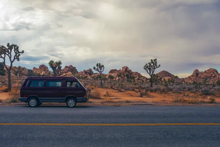 Explore Joshua Tree's desert landscape with an RV rental. A maroon van parked roadside under cloudy skies, surrounded by Joshua trees and rocks.. Volkswagen Westfalia 1991