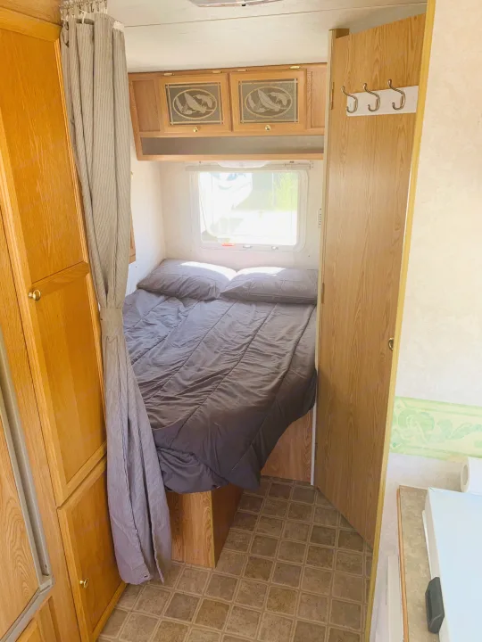 Interior of an RV rental bedroom showing a cozy bed, wooden cabinets, and curtain, perfect for compact travel.. Travel Lite Travel-Lite 800 2003