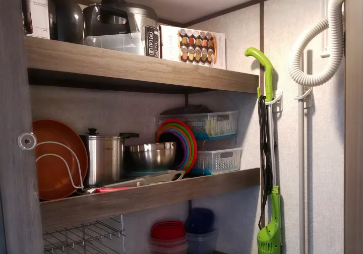 Organized RV rental kitchen shelves hold appliances, cookware, and storage bins. A green vacuum and hose hang on the wall.. Dutchmen Kodiak 2021