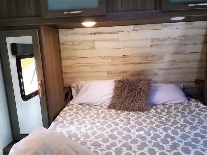 Cozy RV rental bedroom featuring a bed with patterned comforter, fluffy pillow, distressed wood paneling, and window.. Dutchmen Kodiak 2021