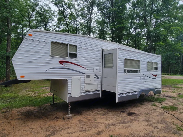 White fifth-wheel RV rental parked in a wooded area, featuring an open door and windows. Ideal travel trailer for camping.. Jayco Eagle 2002