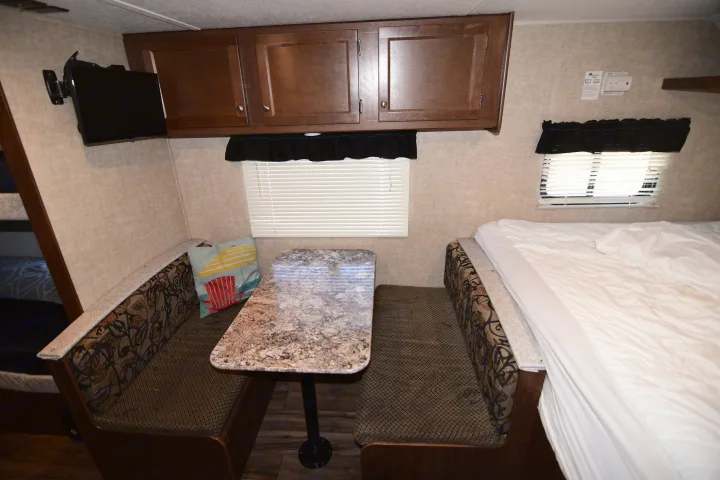 This rv rental features a cozy dinette with a table, two benches, overhead cabinets, a TV, and a comfortable bed on the right.. Keystone RV Hideout 2017