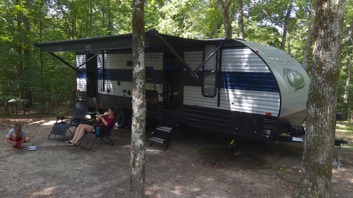 Enjoying an RV rental adventure! A woman relaxes, a child plays with a toy RV by a travel trailer in a shaded campground.. Forest River Cherokee Grey Wolf 2021