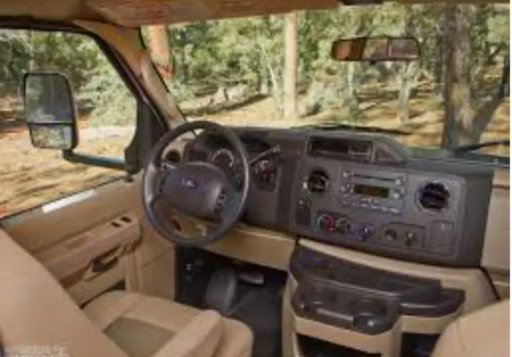 Inside an RV rental: view of the driver's area, including the Ford steering wheel, dashboard, and controls, parked in a forest.. Thor Motor Coach Four Winds Majestic 2013