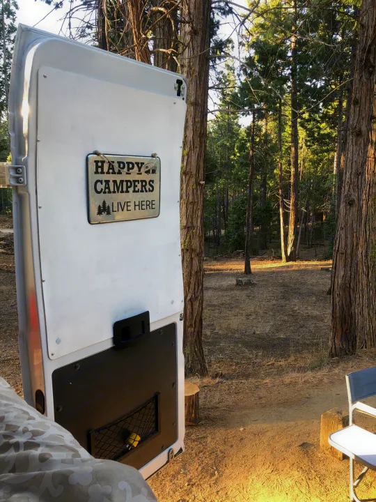 White RV rental door with a 'Happy Campers Live Here' sign, open to a peaceful forest setting with tall trees and dry ground.. dodge promaster 2500 2015