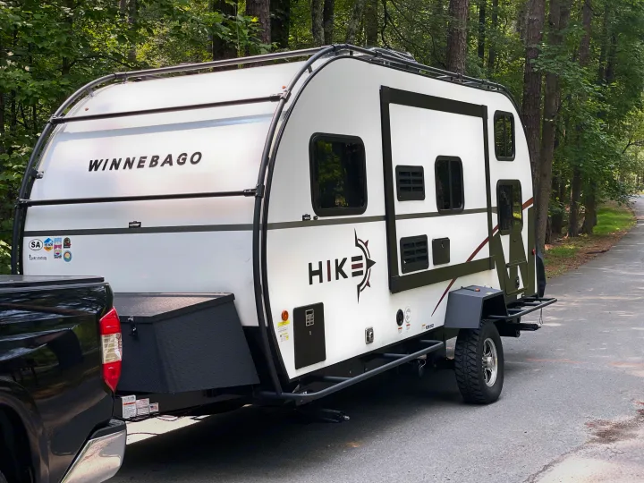 White Winnebago Hike rv rental trailer for outdoor adventures, hitched to a black truck on a wooded road.. Winnebago Hike 2021