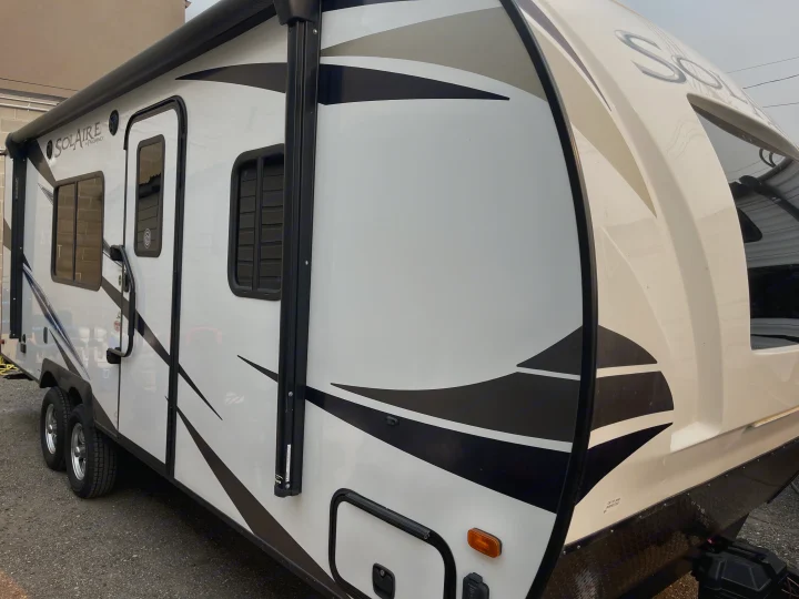 A modern white rv rental travel trailer with black and grey graphics and a side awning, parked on gravel.. Solaire 211BH 2019