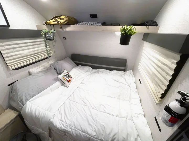 Relaxing RV rental bedroom with a plush white comforter, overhead shelving, pleated window blinds, and charming decor.. Winnebago Hike 2021