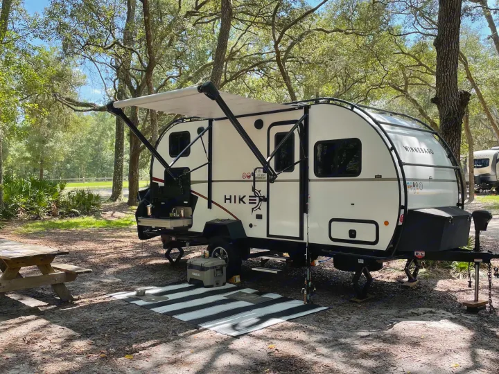 This white Winnebago HIKE rv rental is set up at a shaded campsite. Its awning is out, revealing an outdoor kitchen, cooler, and picnic table.. Winnebago Hike 2021
