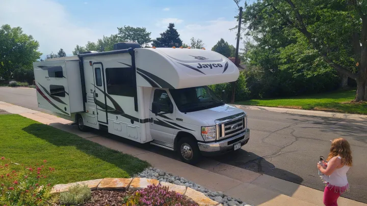 White Jayco Class C RV rental with an extended slide-out, parked on a residential street. A child observes nearby.. Jayco Redhawk 2018
