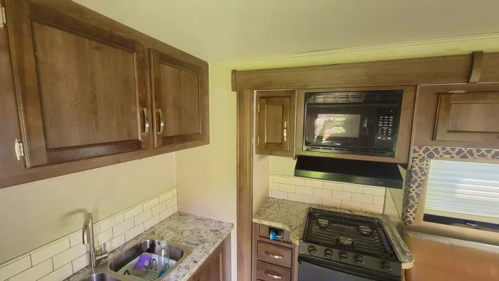 RV rental kitchen with dark wood cabinets, sink, microwave, and stove. Features granite-look counters & subway tile backsplash.. Jayco Redhawk 2018
