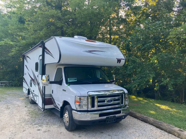 This Winnebago RV rental is parked at a scenic campsite among green trees, ready for your next adventure.. Winnebago Outlook 2020