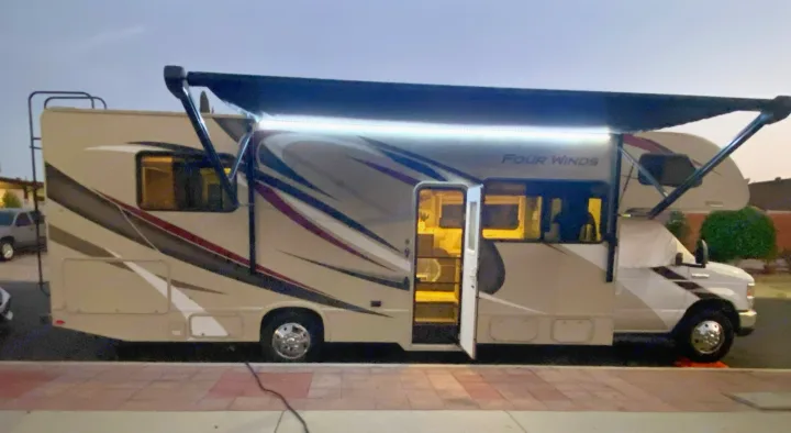 A large, beige Class C RV rental is parked with its door open and an illuminated awning extended, ready for evening adventures.. Thor Four Winds 2020