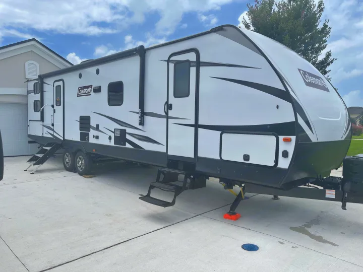 White & black Coleman travel trailer RV rental, parked on a driveway with leveling supports & steps extended, ready for your trip.. Dutchmen Coleman 2021