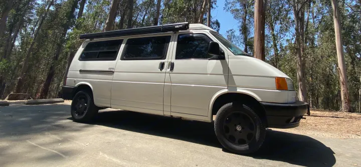 A white classic VW camper van with an awning, parked in a sunny forest. Perfect for your RV rental adventure!. Volkswagen T4 Multivan Westfalia 1995