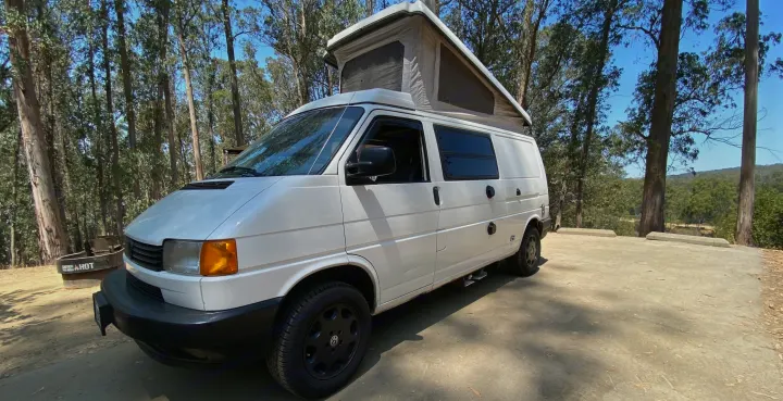 This white pop-top camper van, perfect for an RV rental adventure, is parked in a sunny forest.. Volkswagen T4 Multivan Westfalia 1995