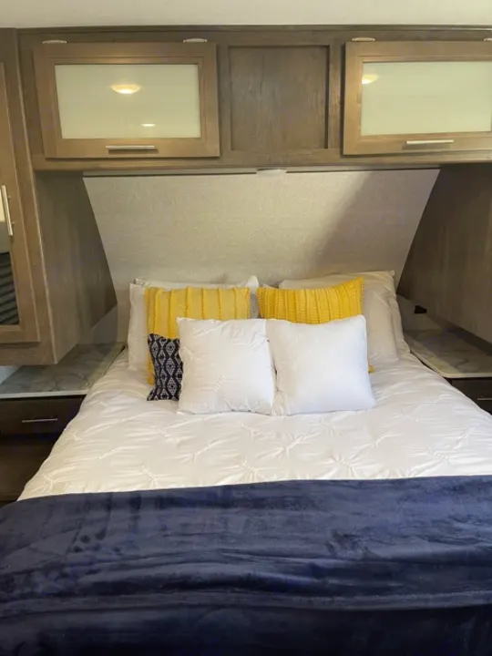 Cozy RV rental bedroom with white bedding, yellow pillows, a blue blanket, and built-in cabinets.. Dutchmen Coleman 2021