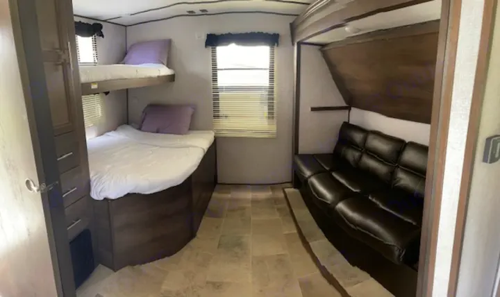 RV rental interior featuring comfortable bunk beds with purple pillows, a brown leather sofa, and a bright window.. Dutchmen Coleman 2021