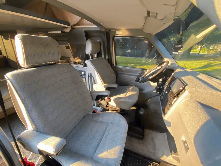 Interior of an **RV rental** camper van, featuring sunlit front seats, steering wheel, dashboard, and a compact kitchen.. Volkswagen T4 Multivan Westfalia 1995